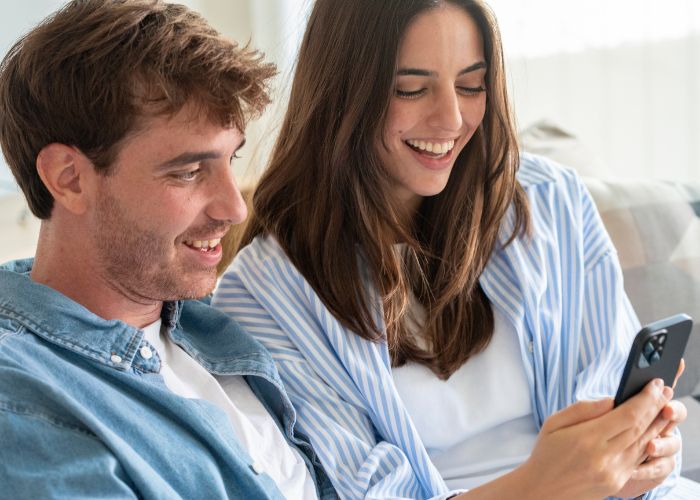 Smiling Couple Sitting On A Sofa And Looking At Photos On A Smartphone.