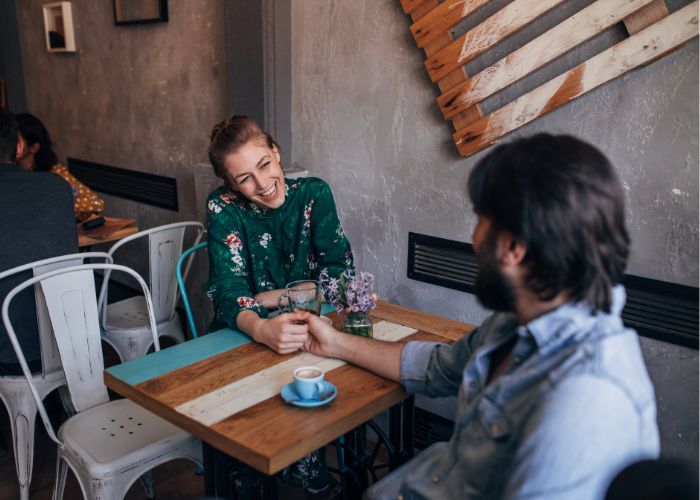 Two People Smiling And Holding Hands Across A Café Table During A Relaxed Coffee Date