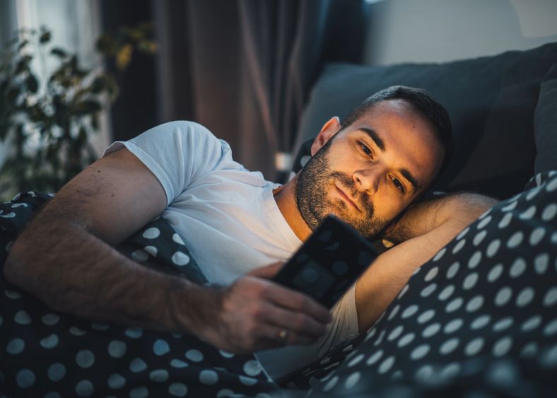 Man Lying In Bed At Night, Smiling While Looking At His Phone