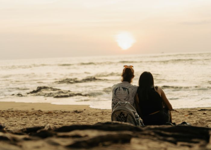 Two People Sitting On The Beach Watching The Sunset