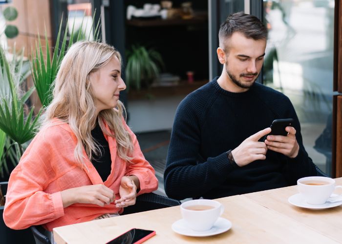 Couple Having Meeting And Man Using Smartphone In Outdoor Cafe