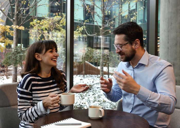 Woman And Man Chatting Over Coffee At A Café Table By Large Windows.