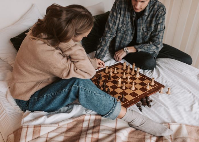 Couple Sitting On A Bed And Playing Chess Together.
