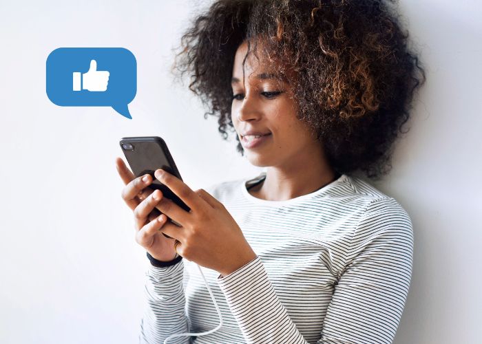 A Woman Smiling At Her Phone With A Like Emoji Bubble