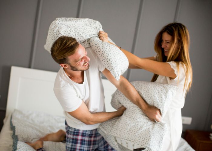 Laughing Couple Having A Playful Pillow Fight On Their Bed.