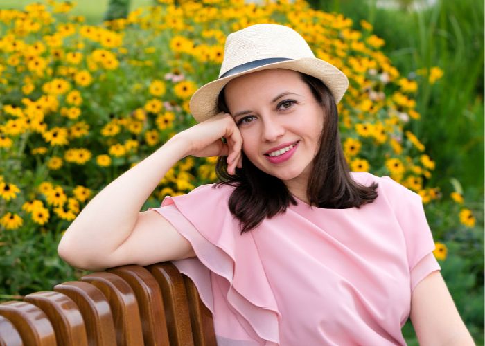 Woman In A Pink Dress And Hat Smiling While Sitting On A Bench