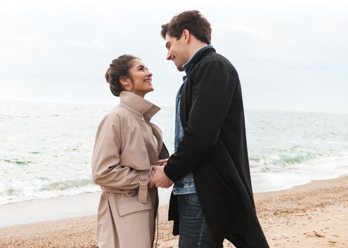 A Happy Couple Holding Hands On A Beach, Smiling At Each Other By The Ocean.