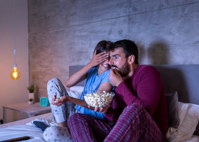 Couple In Pajamas Sharing Popcorn While Watching A Scary Movie Together In Bed